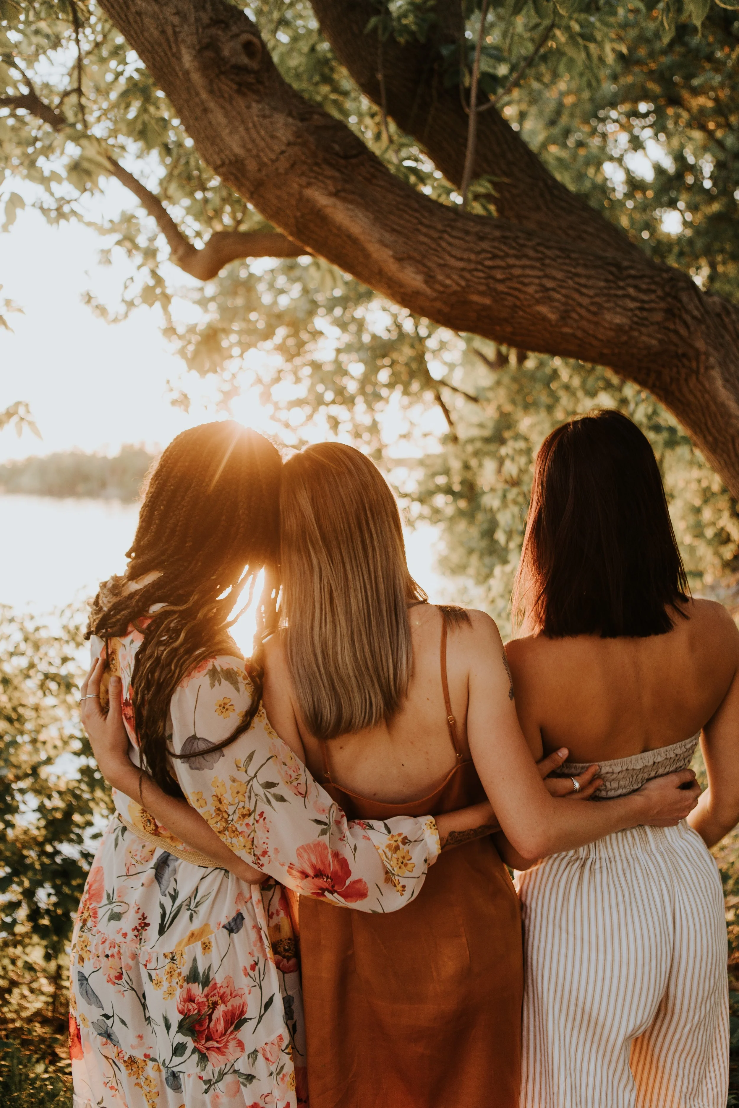 Three women standing close together under a tree, facing a body of water at sunset.