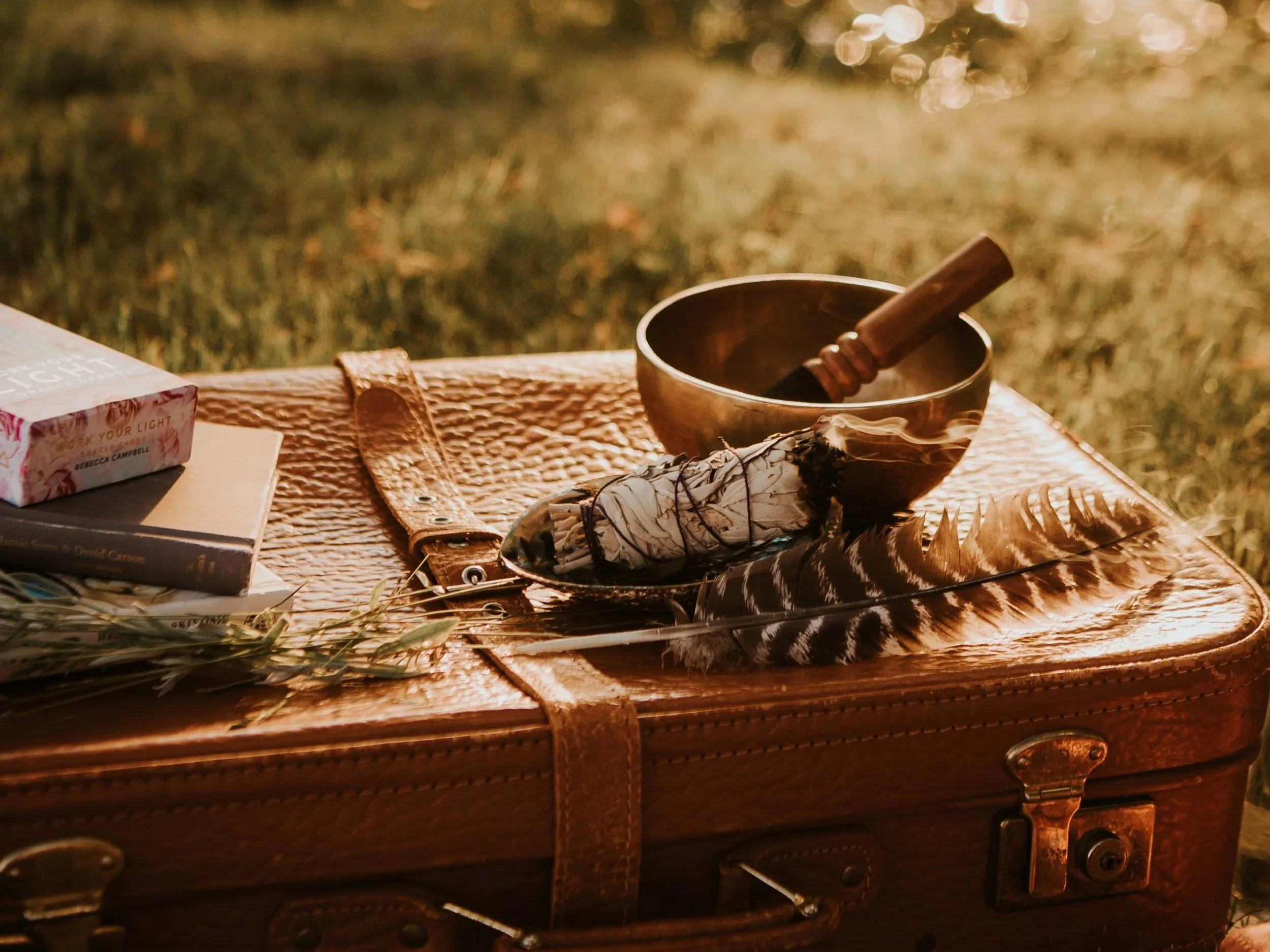 A leather suitcase on the grass with a snack, books, an herb bundle, a feather, a bowl, a stick, and a burning smudge stick on top, in warm sunlight.