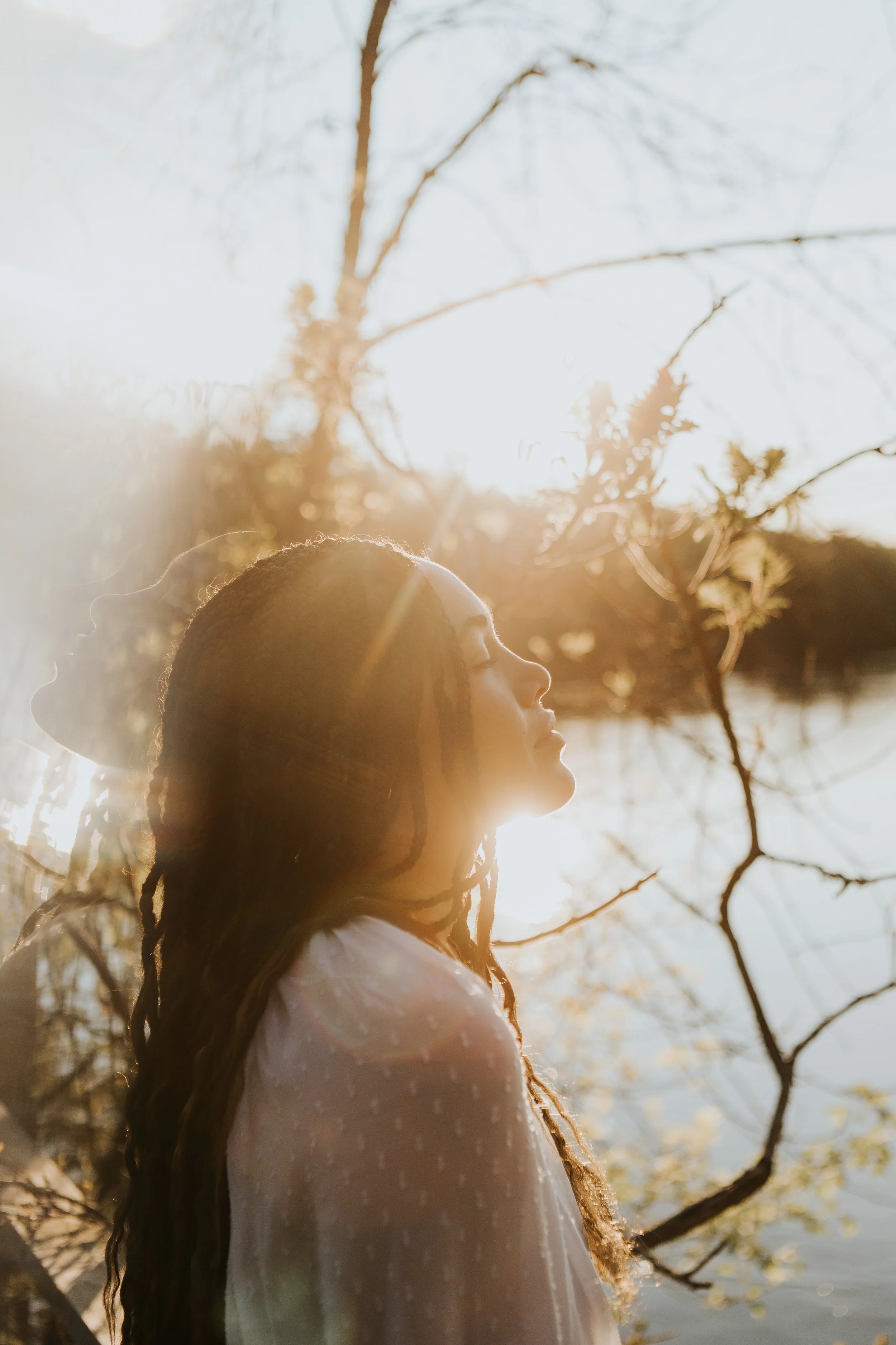 A woman with long braids, wearing a white dress with puffed sleeves, stands outdoors with her eyes closed as the sun rises or sets behind her, creating a warm glow and lens flare, with leafless tree branches visible.