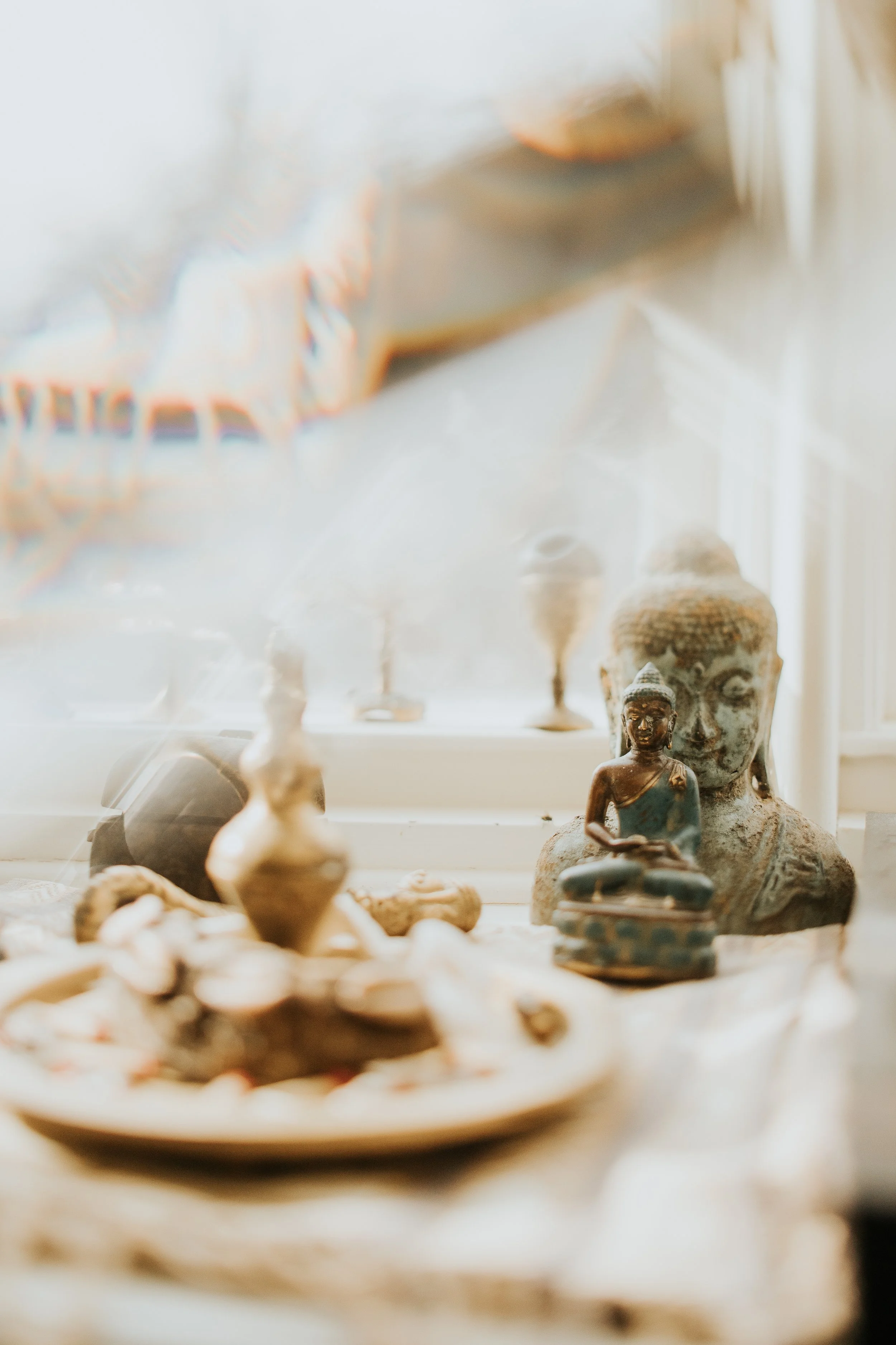 Close-up of decorative Buddha statues and small artifacts on a wooden surface near a window, with a blurred colorful cloth in the background.