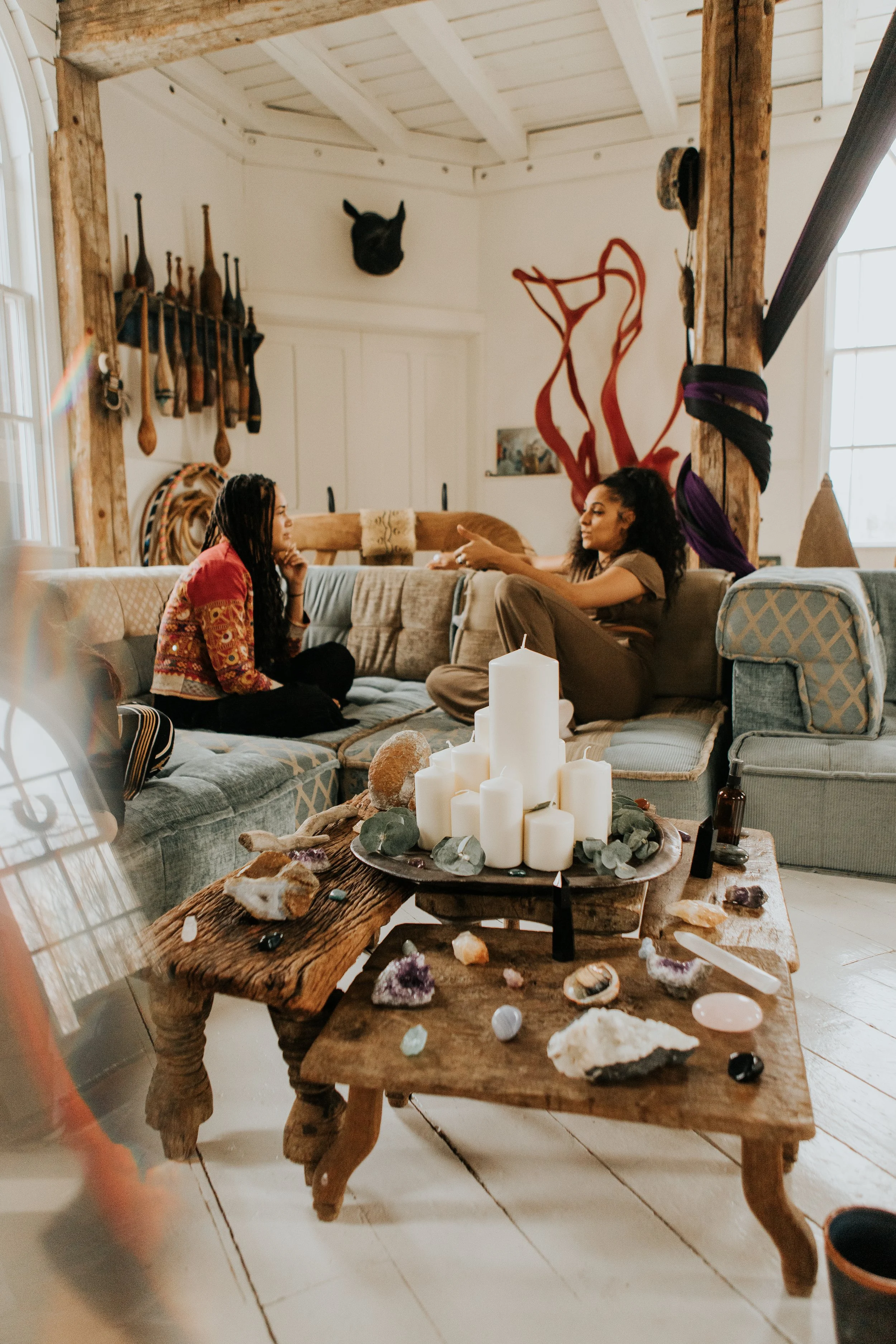 Two women sitting on a couch in a cozy room with decorated wooden walls and a high, white ceiling. They are engaged in conversation. The room has a rustic style with candles and crystals on a wooden coffee table in front of them.
