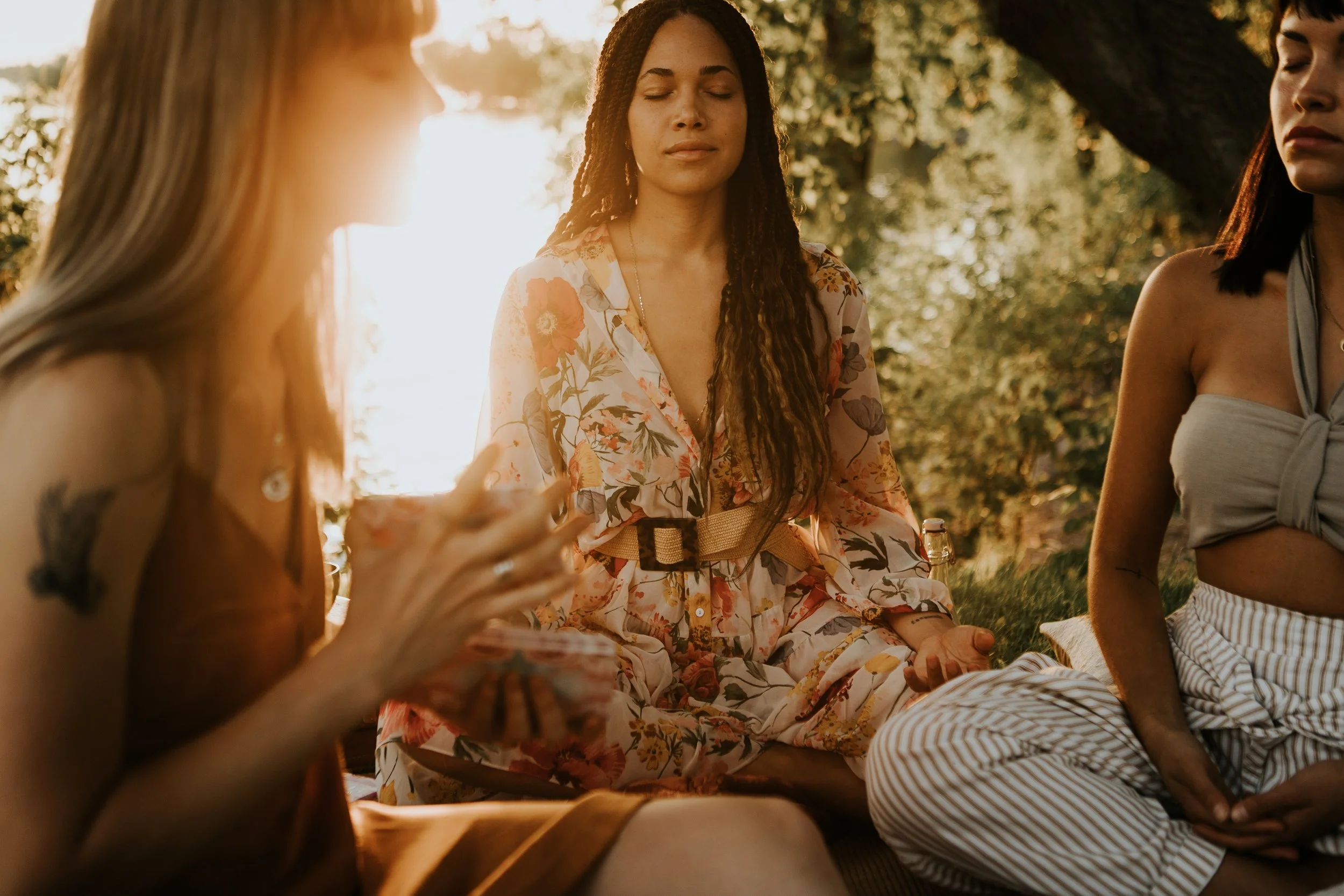 Three women sitting outdoors during sunset, engaged in meditation or mindfulness, with a large tree and greenery in the background.
