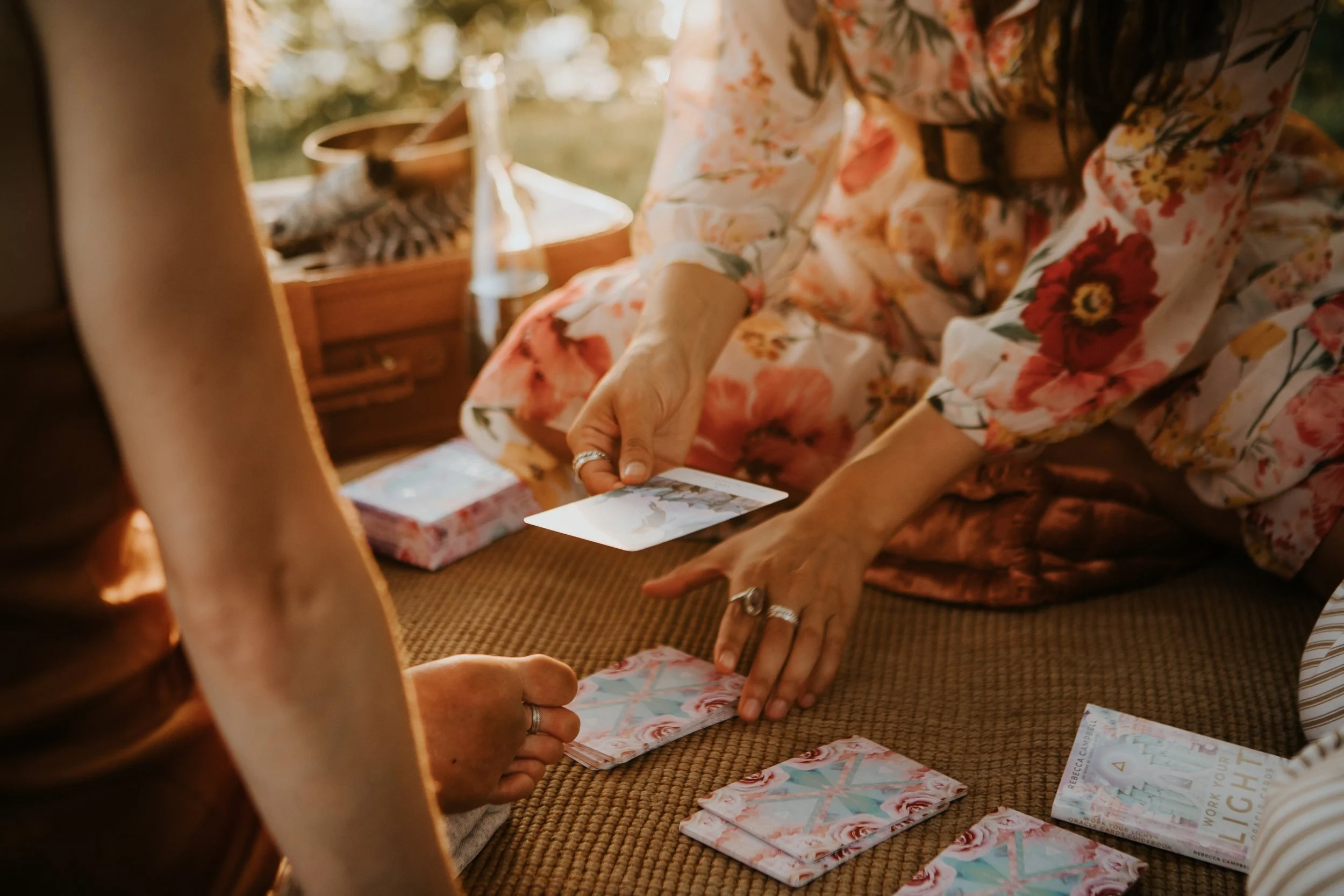 Two people are playing tarot card game on a brown mat outdoors. One person is handing a card to the other. They are wearing rings and a floral dress. There are tarot cards and a book on the ground.