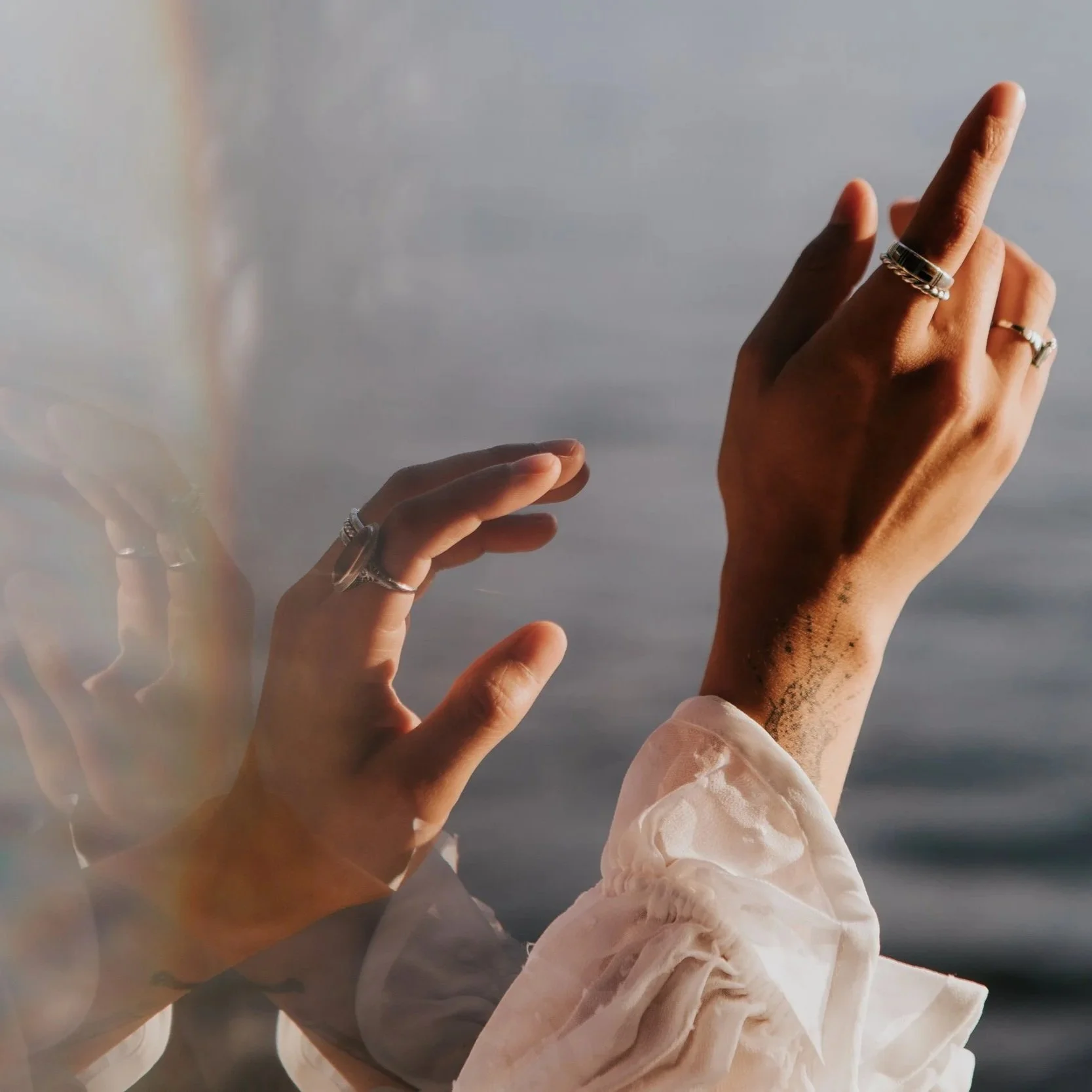 Close-up of two hands with rings reaching towards the sky with water in the background.