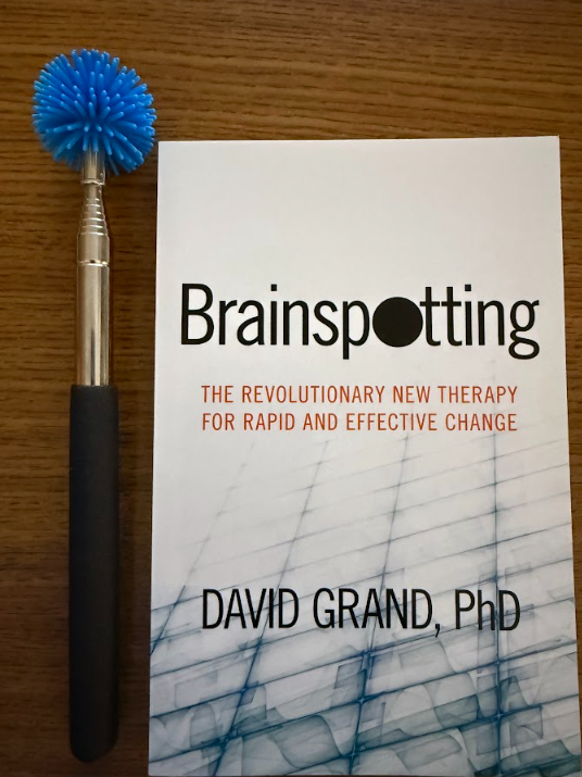 A book titled "Brainspotting" by David Grand, PhD, placed on a wooden surface next to a handheld tool with a blue spiky head.