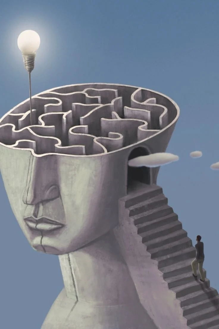 A surreal sculpture of a human head with a maze inside the top, resembling a brain. There is a staircase leading into the head's side, with a person sitting at the bottom, and a lit light bulb balanced on a pin on the maze.