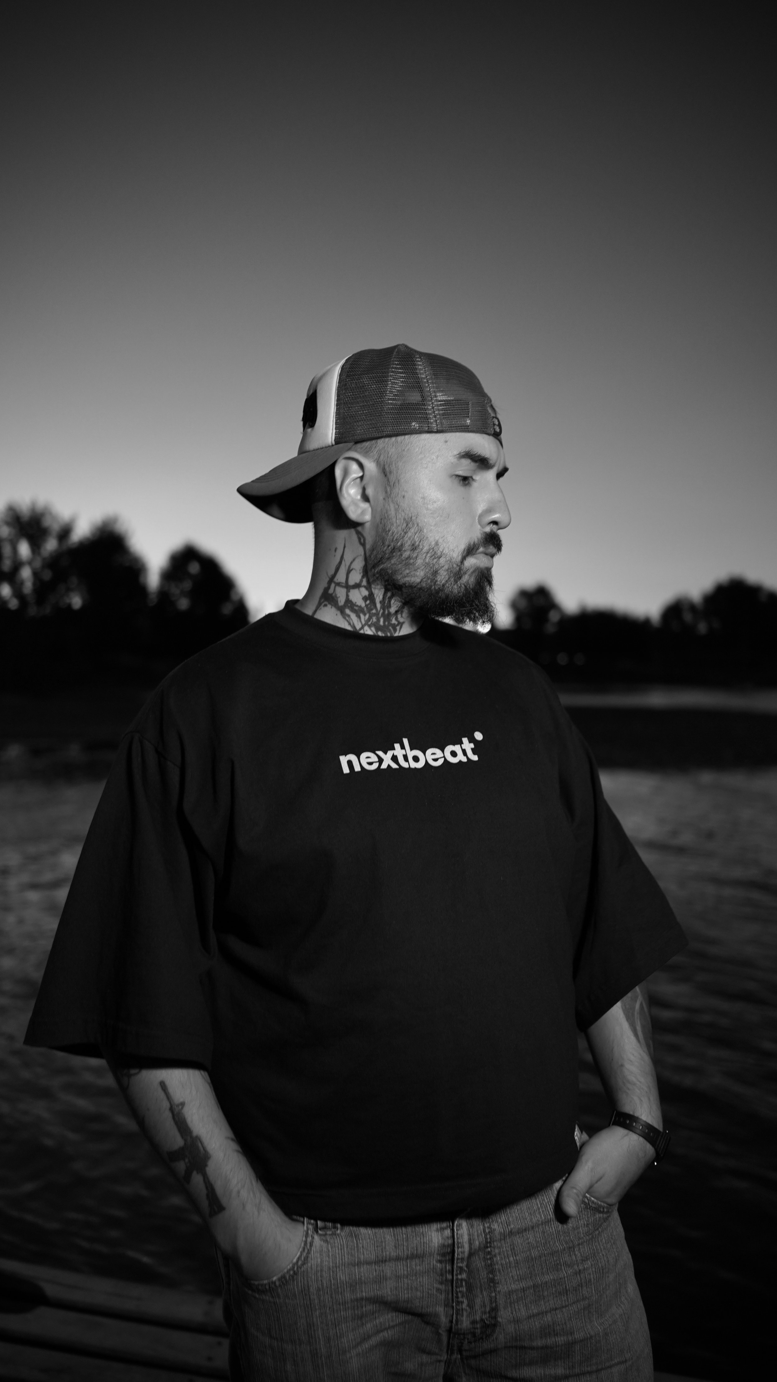 Joven con barba y tattoos, lleva una gorra de béisbol y camiseta negra con la palabra 'nextbeat' en una zona al aire libre durante el atardecer o amanecer, con fondo de árboles y cielo en escala de gris.