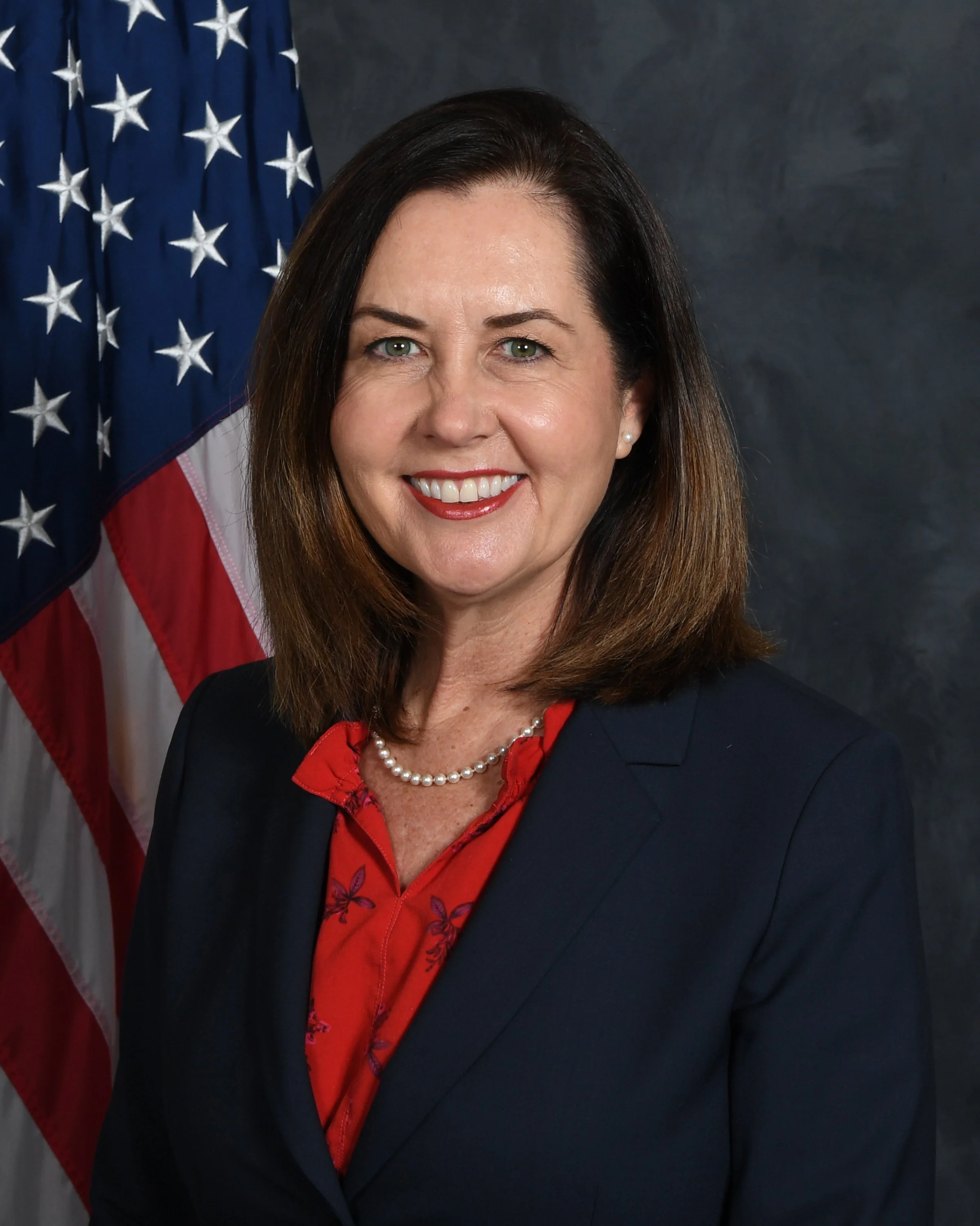 Portrait of Kim Carr with dark brown hair, green eyes,  and pearl earrings, smiling in front of an American flag.