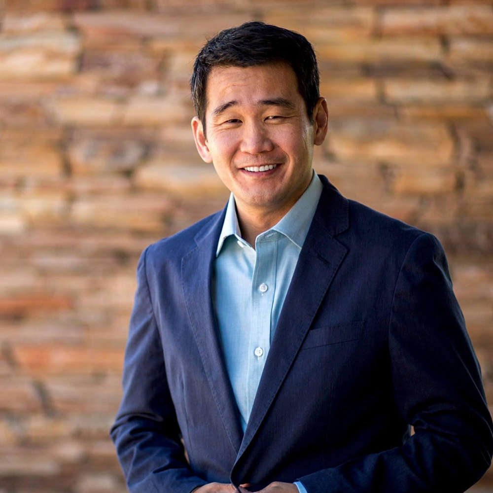 Rep. Dave Min in a navy blazer and light blue shirt smiling against a wooden background.