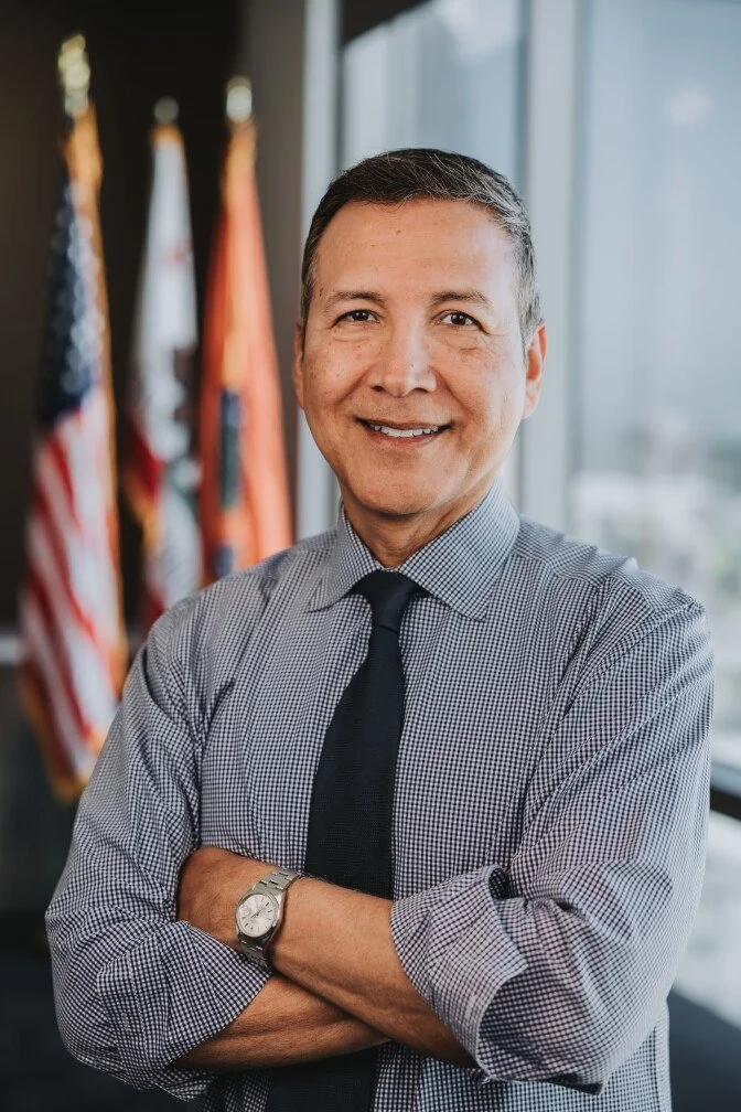 Supervisor Vince Sarmiento in a checkered shirt and black tie, is standing with his arms crossed in front of a window with American flags in the background.