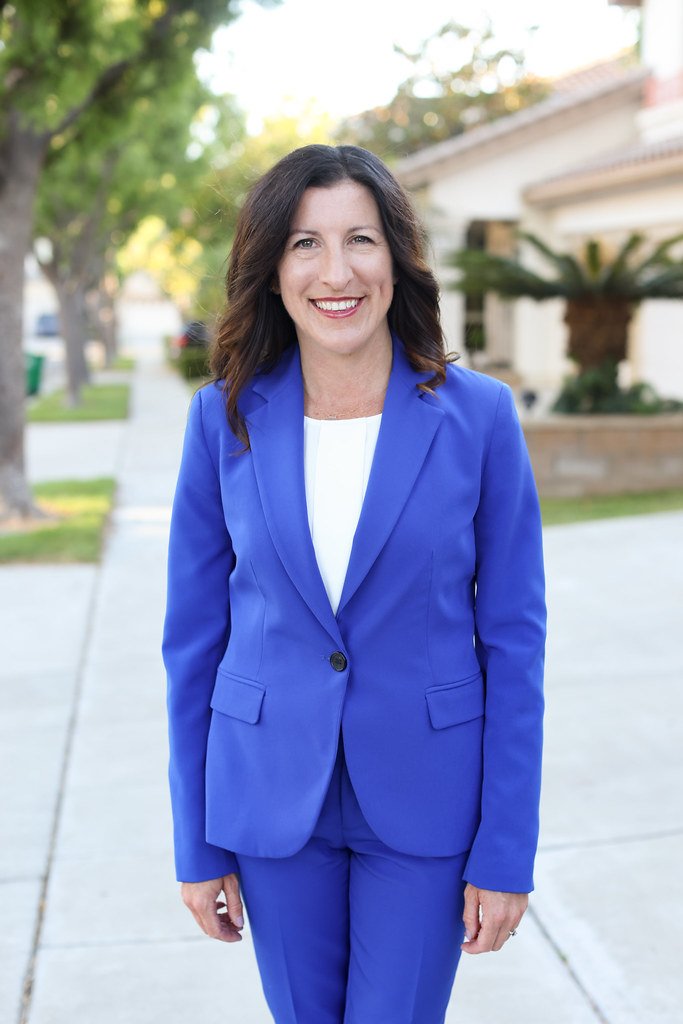 Assembly Member Cottie Petrie-Norris in a bright blue suit is smiling outdoors on a sidewalk with trees and houses in the background.