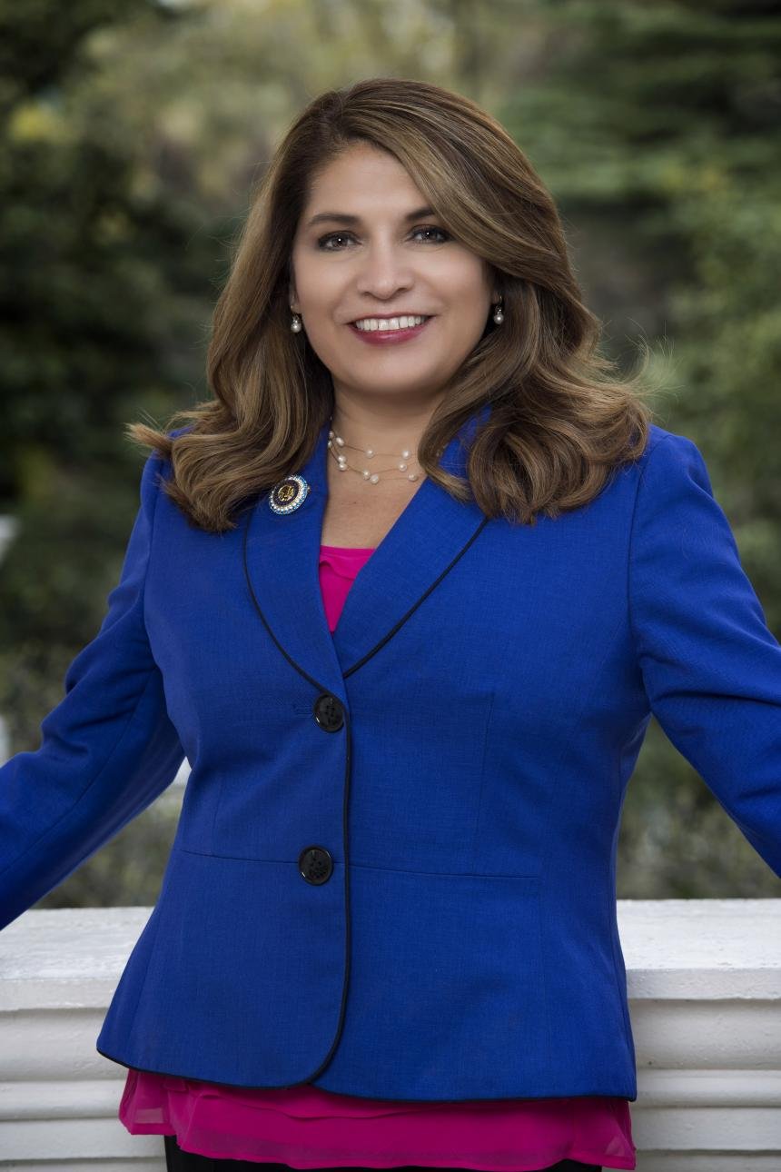 Assembly Member Sharon Quirk-Silva is smiling, wearing a royal blue blazer, pink top, pearl necklace, and earrings, standing outdoors with blurred green foliage in the background.