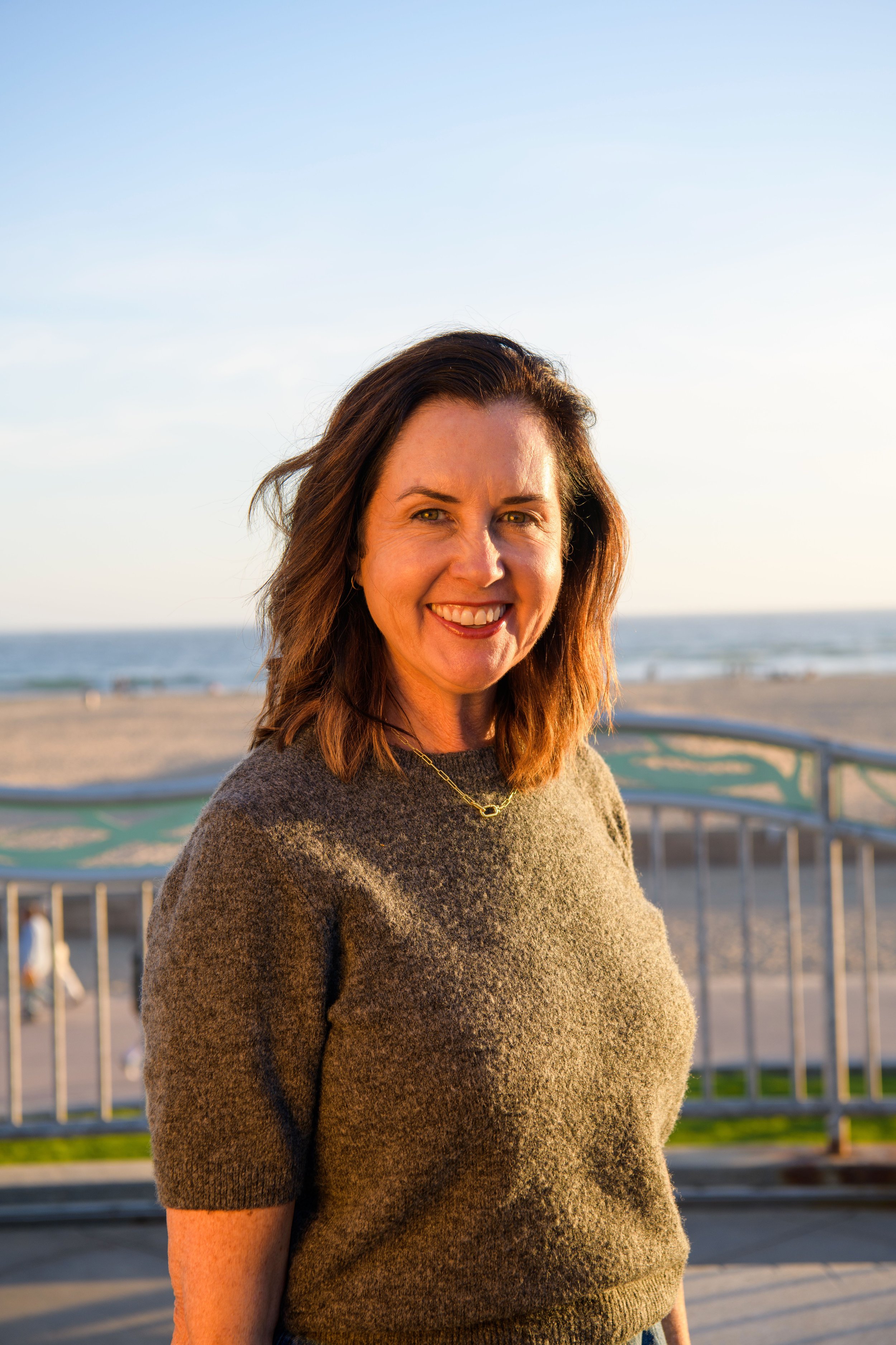 Kim Carr is smiling on the beach during sunset, standing near a railing with the ocean in the background.