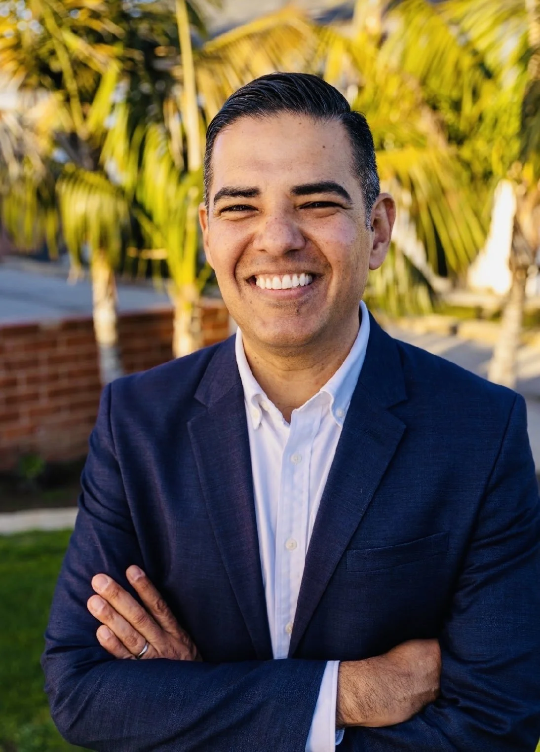Rep. Robert Garcia  is smiling with arms crossed, wearing a dark blue suit and white shirt, standing outdoors with palm trees and a brick wall in the background during daytime.
