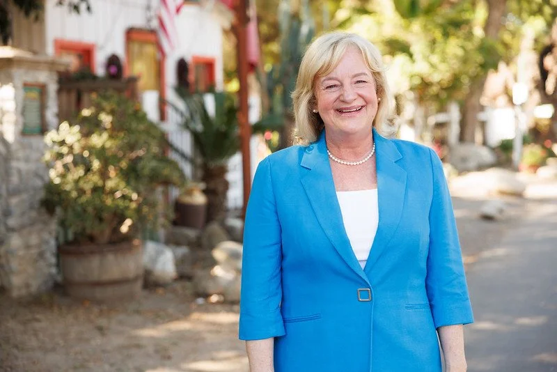Supervisor Katiran Foley is wearing a bright blue blazer and white top, standing outdoors on a sunny day with a colorful, rustic village background.