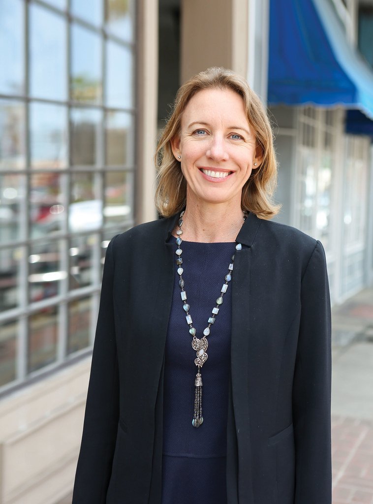 Senator Catherine Blakespear is smiling outdoors in front of a storefront window, wearing a black blazer and a long necklace.