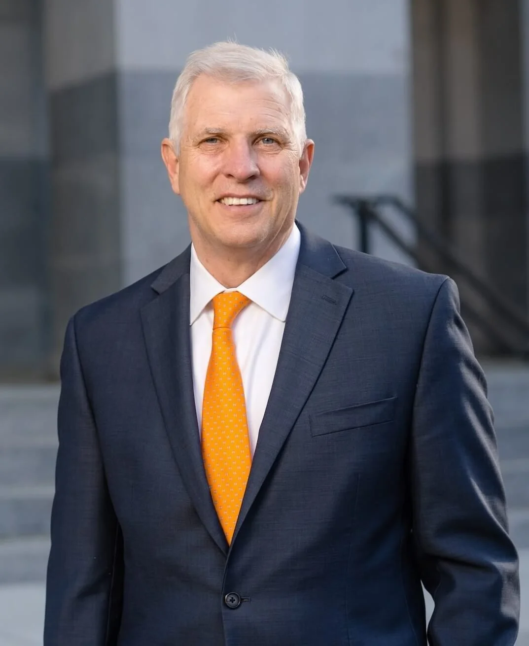 Senator Tom Umberg has gray hair, he's smiling, and wearing a dark suit, white shirt, and orange tie. He is standing outdoors in front of gray stone building steps.