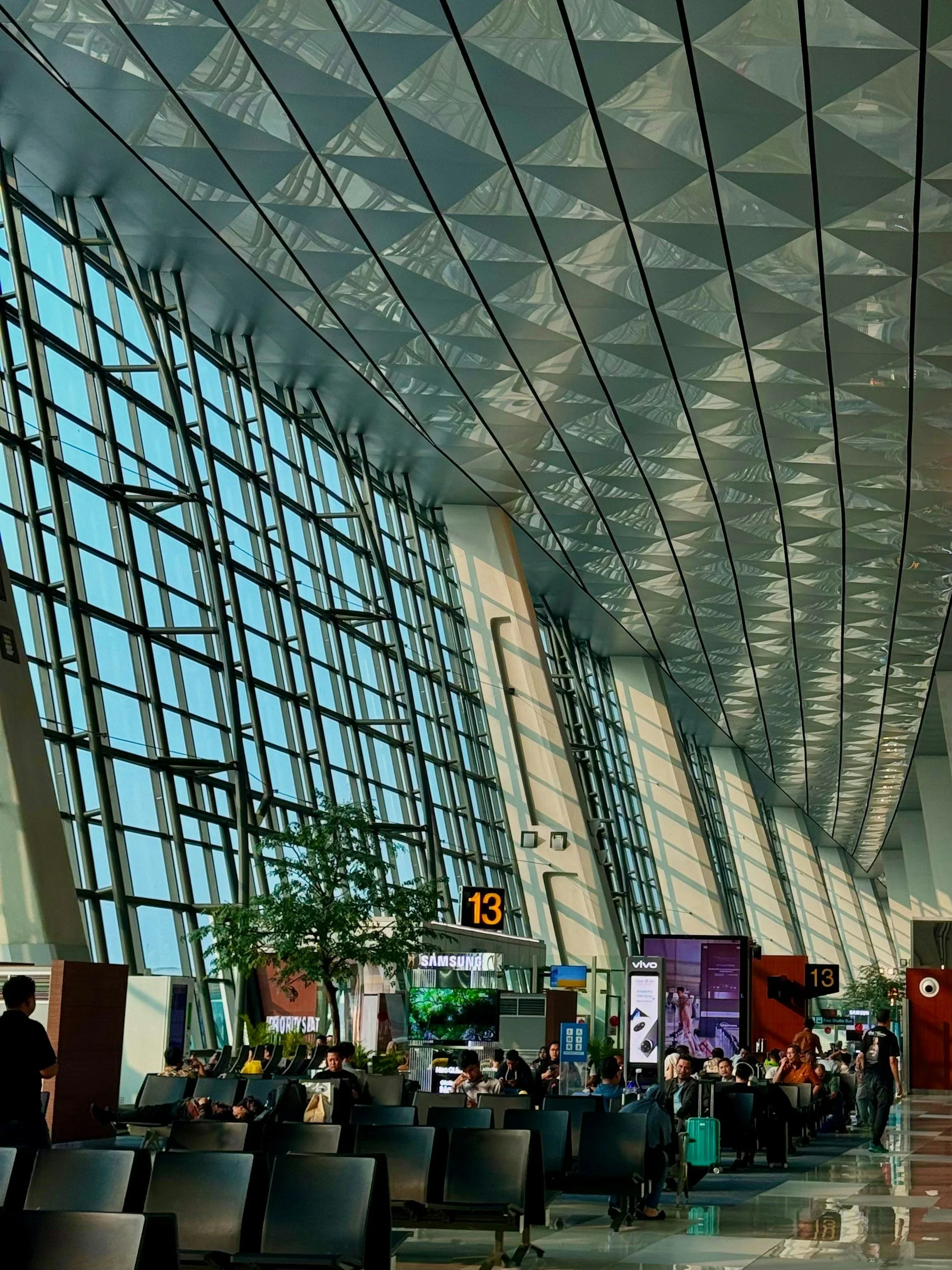 Interior of an airport terminal with high glass windows, metal ceiling, and people waiting with luggage. Digital flight information display shows gate 13.