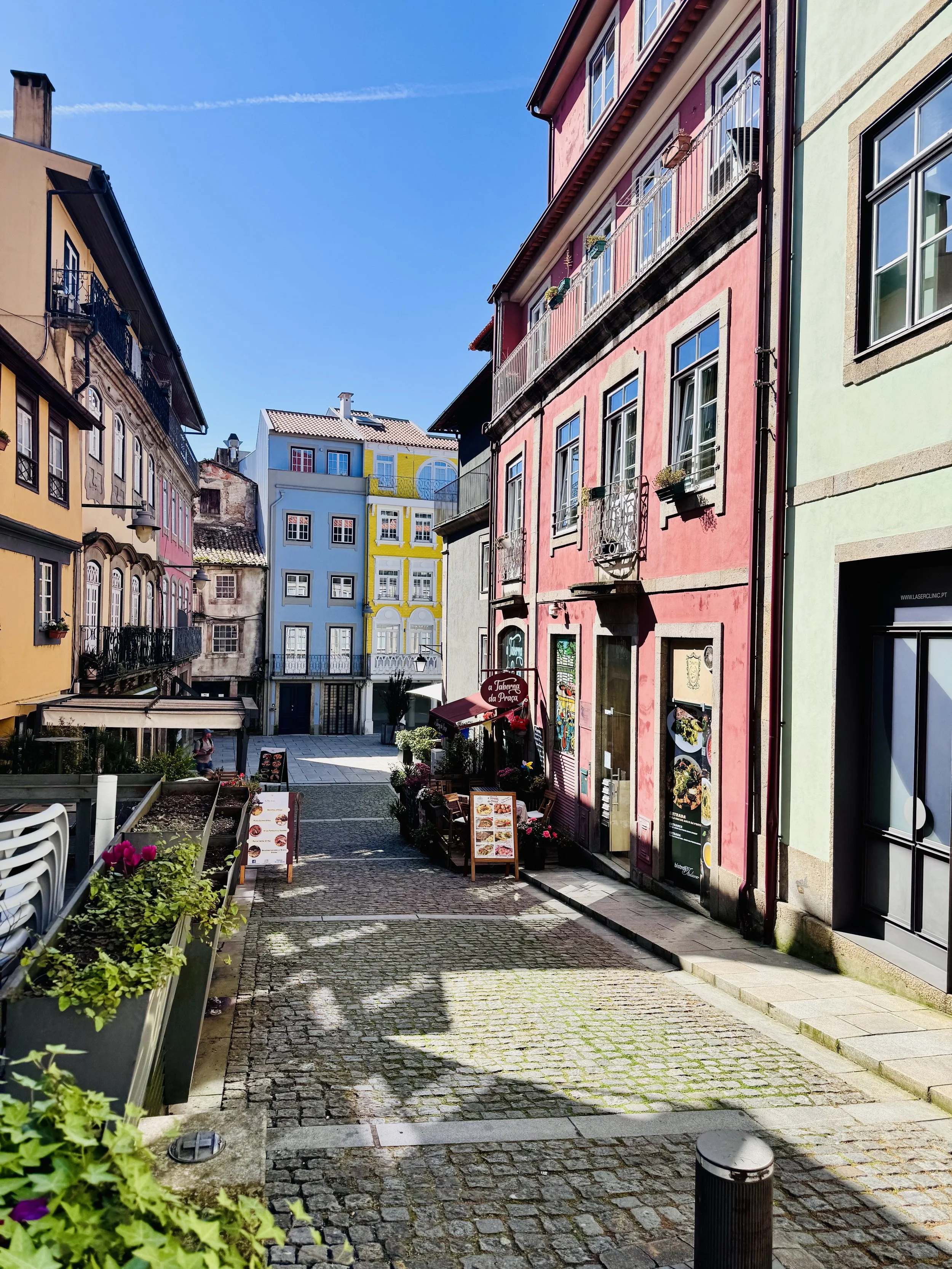 Colorful buildings on a European street with cobblestone pavement, flowers in planters, and a clear blue sky.