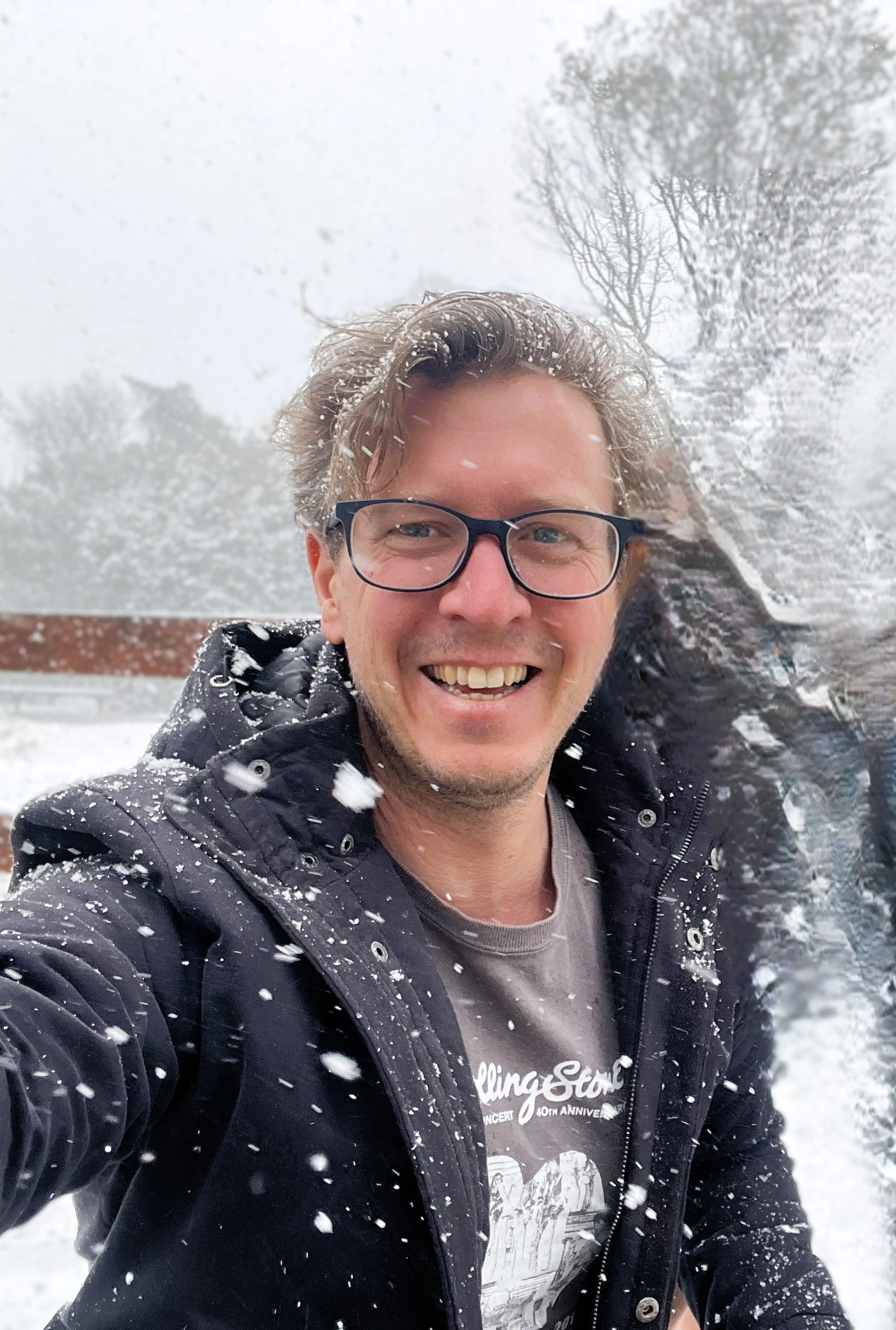 A smiling man with glasses taking a selfie in a snowy outdoor setting, snow falling around him and snow on his jacket.