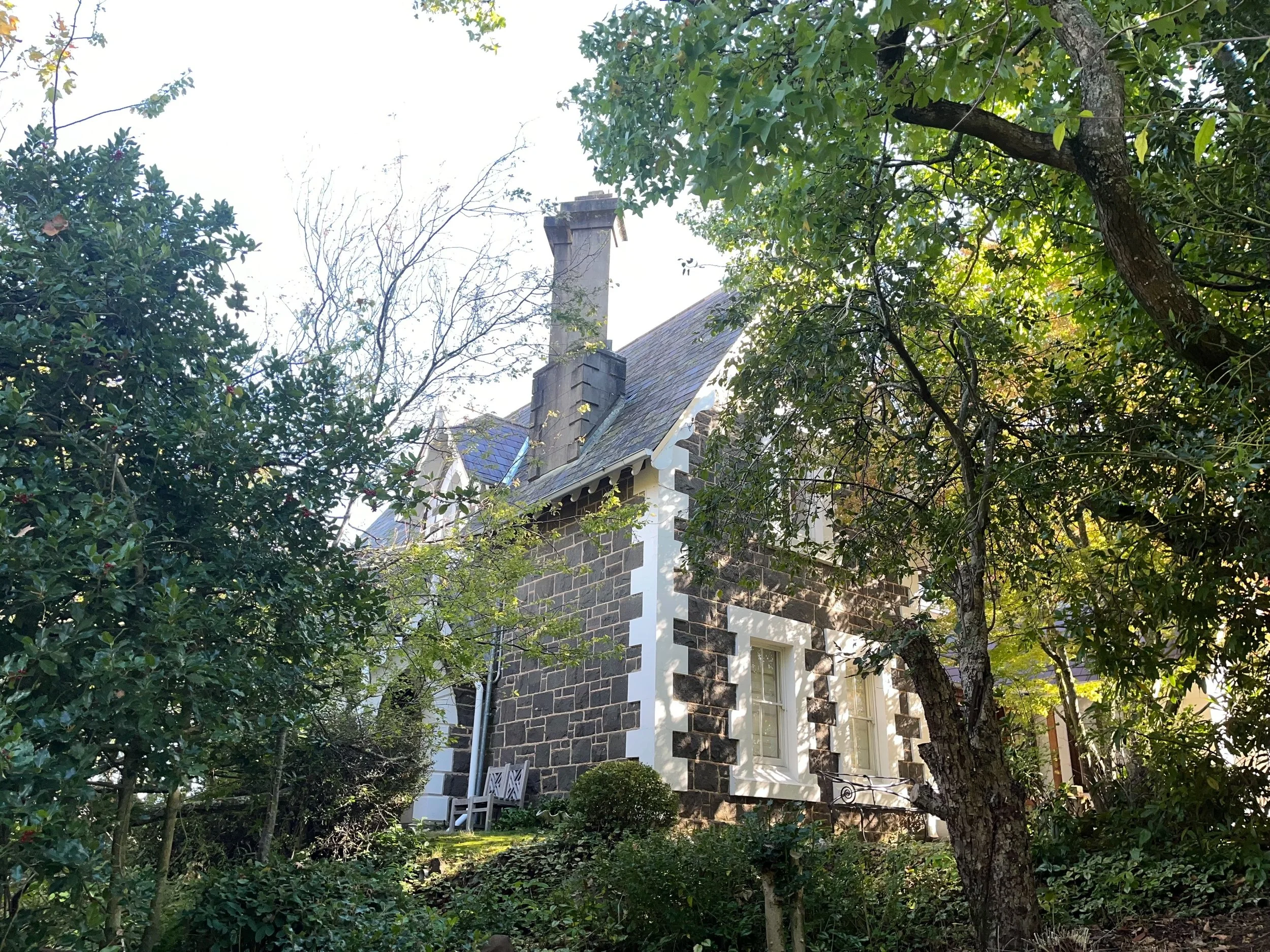 A charming house made of dark bricks with white trim, situated behind lush green trees and bushes, with a bench visible outside.