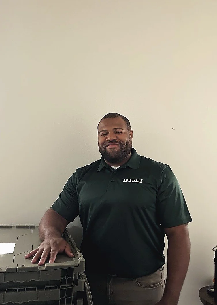 A man with a beard wearing a black polo shirt with a logo that reads 'TOTEFLEET' stands next to a stack of gray plastic storage bins in front of a plain light-colored wall.