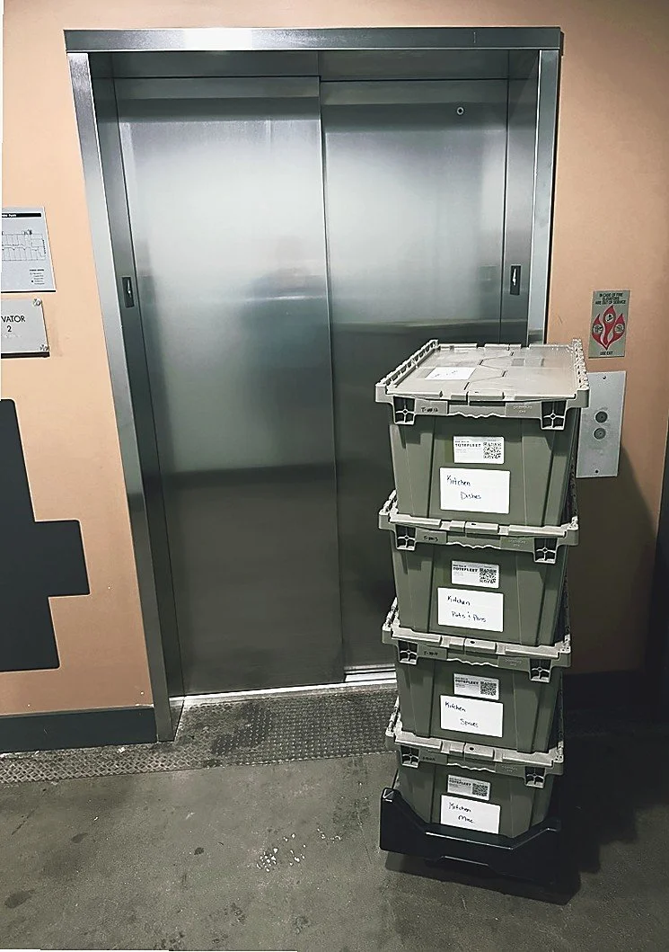 A stack of labeled plastic storage bins next to an elevator door in a building, with labels indicating they contain kitchen dishes, pots and pans, spices, and toilet paper.