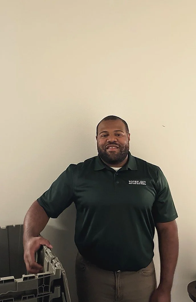 Man standing against a plain wall, wearing a dark green polo shirt with a logo, holding a plastic storage bin.