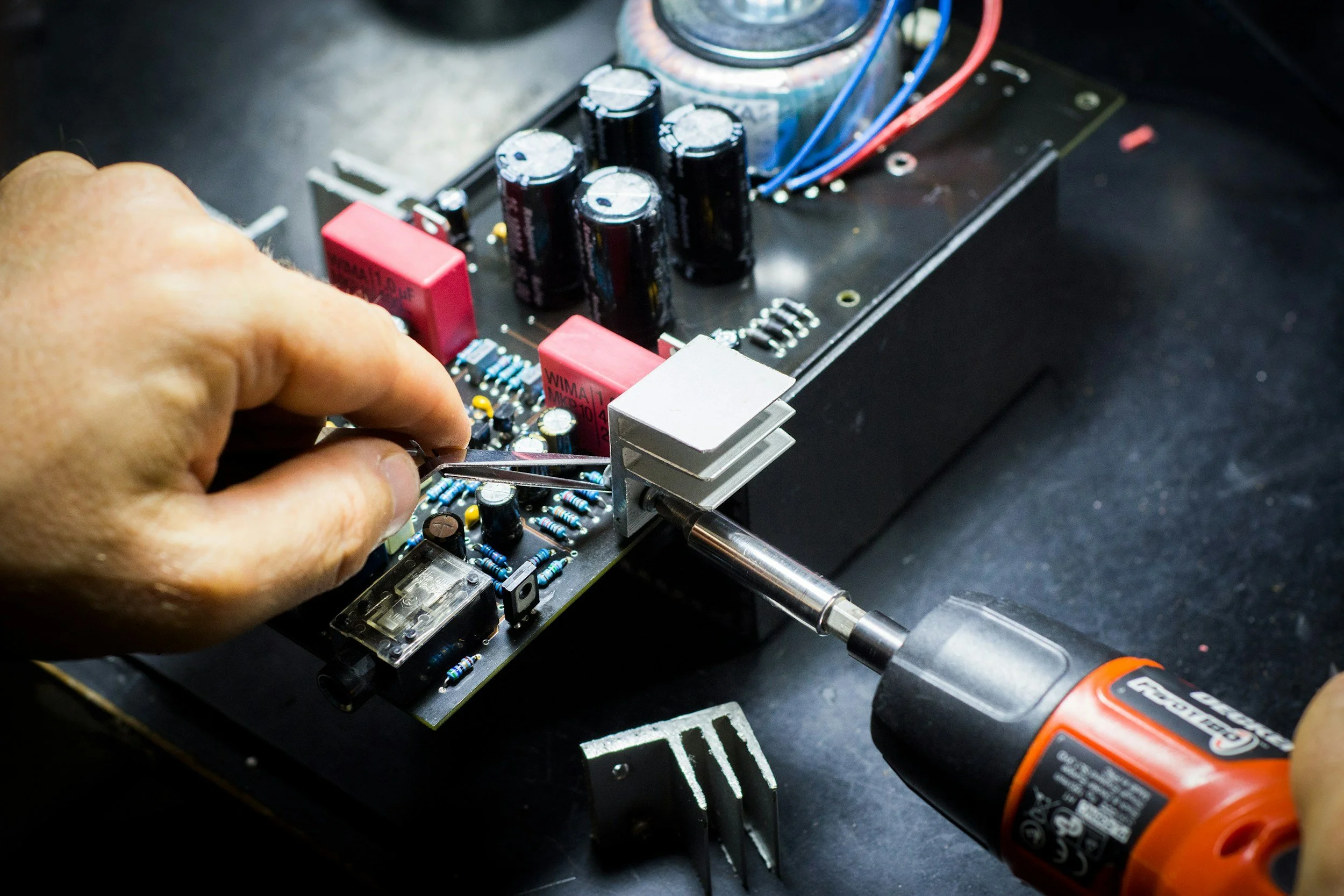 A person's hand using a screwdriver to work on an electronic circuit board with various components, including capacitors, resistors, and a transformer.