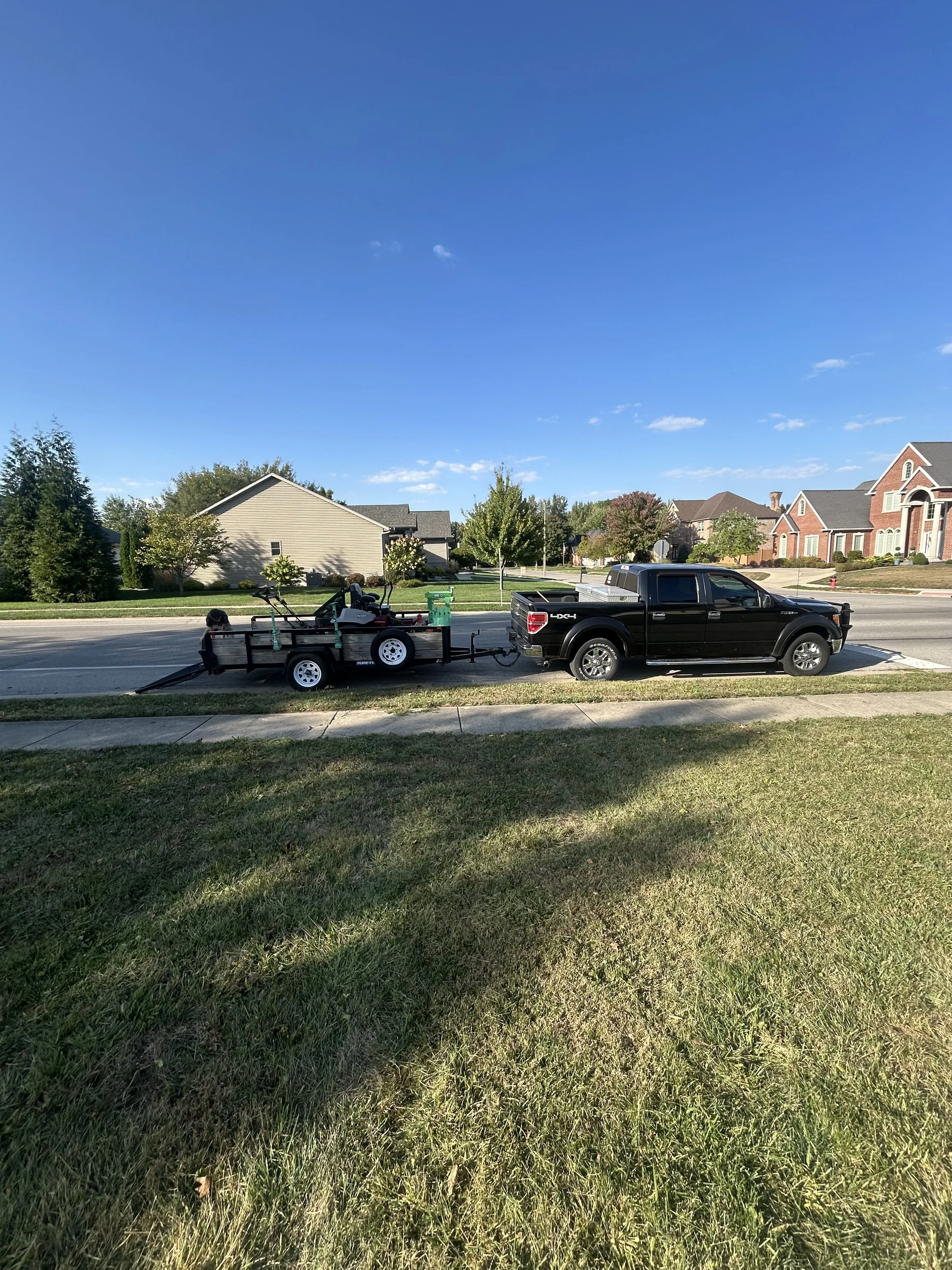 A black pickup truck towing a small trailer with equipment and a bicycle, parked on a suburban street with houses and trees under a clear blue sky.
