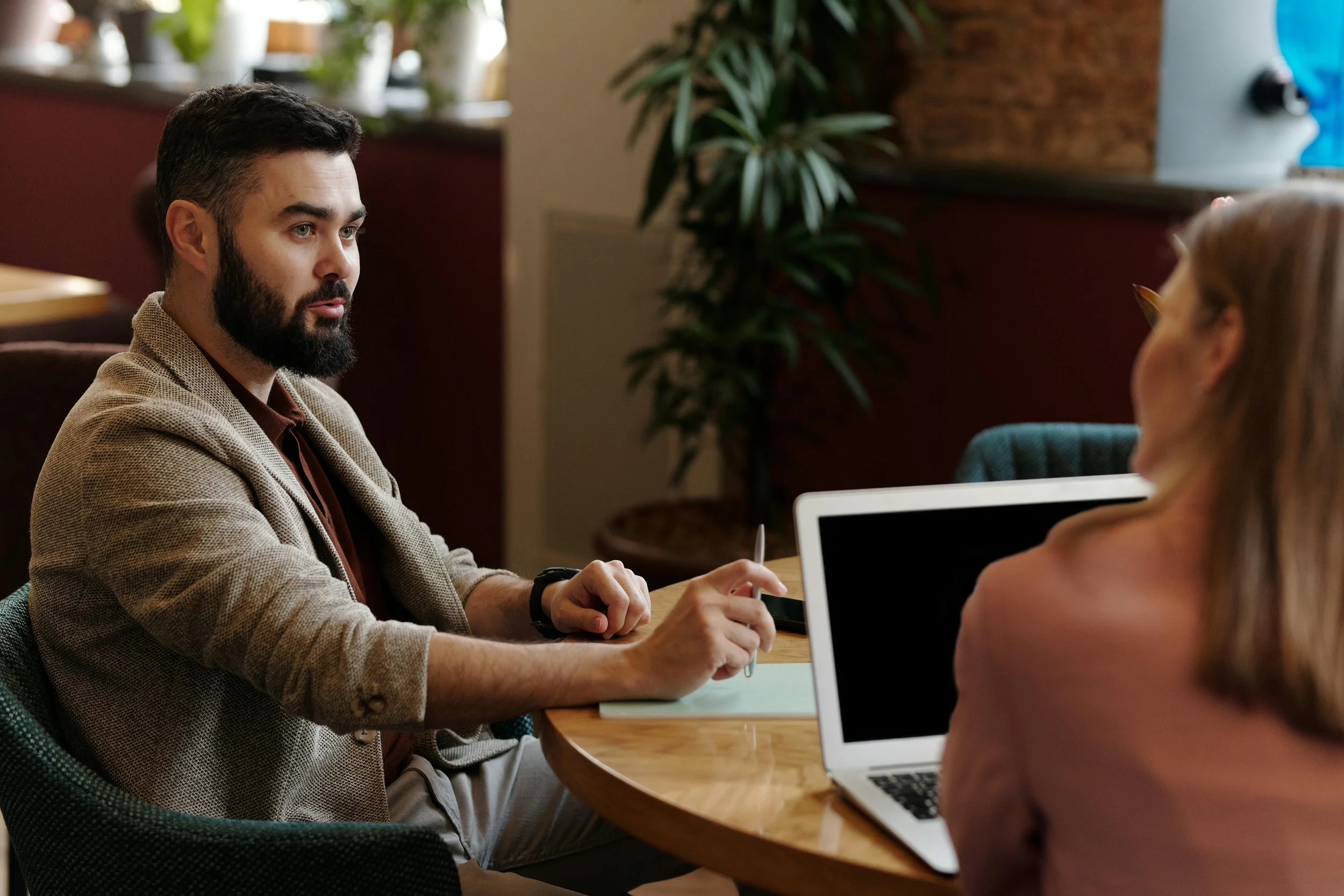 A man with a beard and a beige blazer sitting at a table having a conversation with a woman with glasses at an indoor cafe or restaurant.