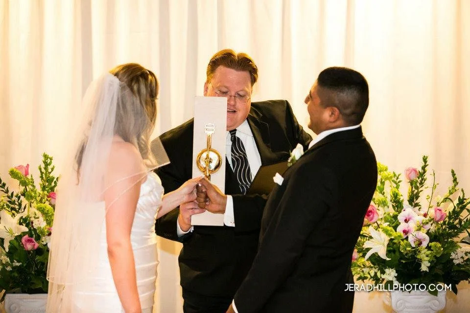 A bride and groom holding hands during their wedding ceremony, with an officiant in the middle, in front of floral arrangements.