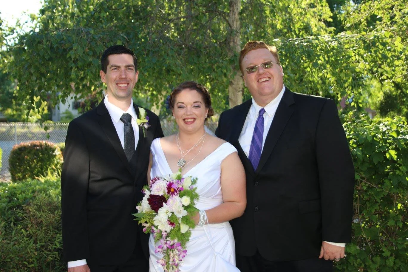 A bride, a groom, and their officiant standing outdoors in a garden, all smiling, with trees and bushes in the background.