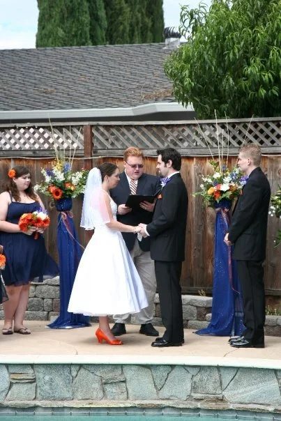 Bride and groom exchange vows during outdoor wedding ceremony with bridesmaids and groomsmen in formal attire, decorated with flower arrangements and draped fabric.