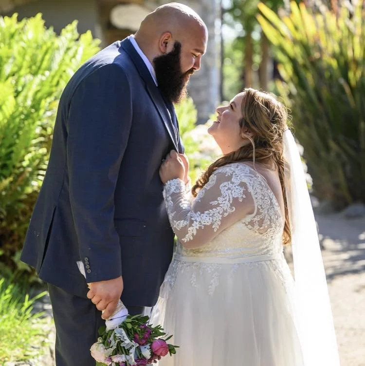A man and woman in wedding attire standing close outdoors, gazing at each other, with the woman holding a bouquet of flowers.
