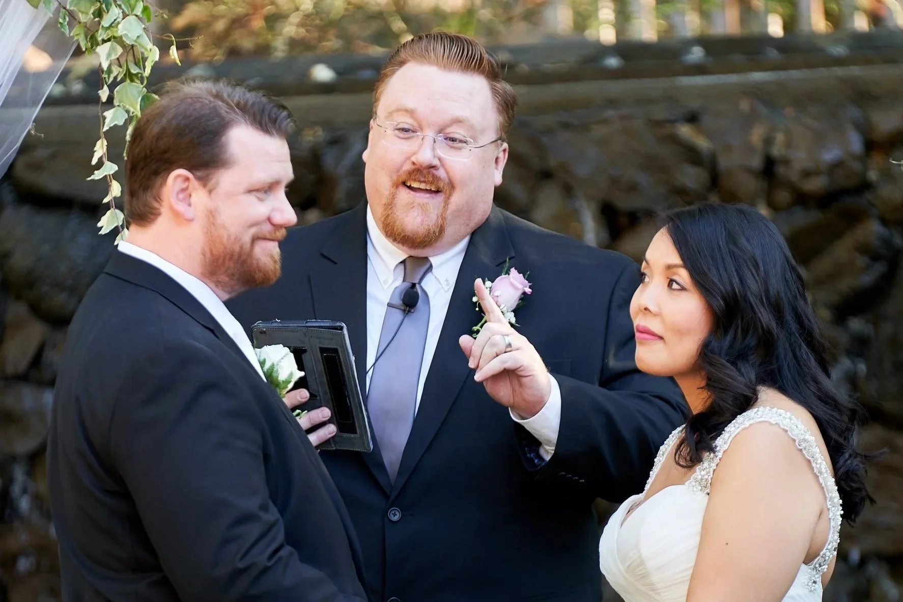A wedding ceremony outdoors with a officiant, a groom, and a bride. The officiant is speaking and pointing his finger, while the groom and bride listen. The groom has a beard and reddish hair, and the bride has long black hair. The background features rocks and greenery.