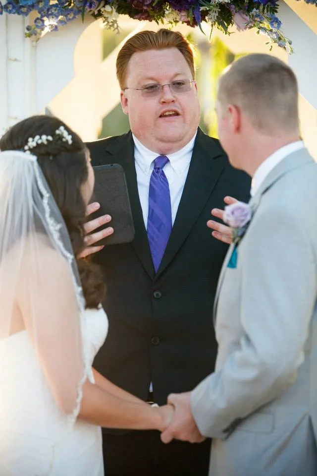 A wedding ceremony with an officiant presiding over a couple holding hands and exchanging vows outdoors.
