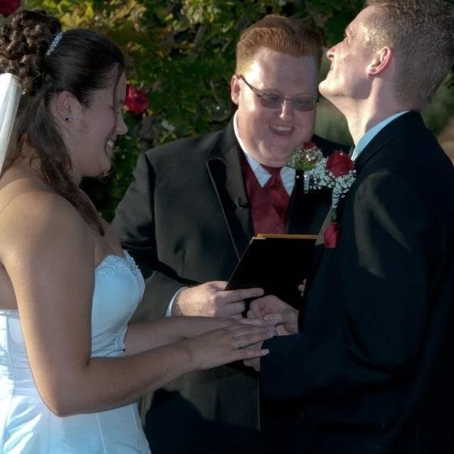 A wedding ceremony with a bride and groom exchanging rings, officiant reading from a book, outdoors with greenery.