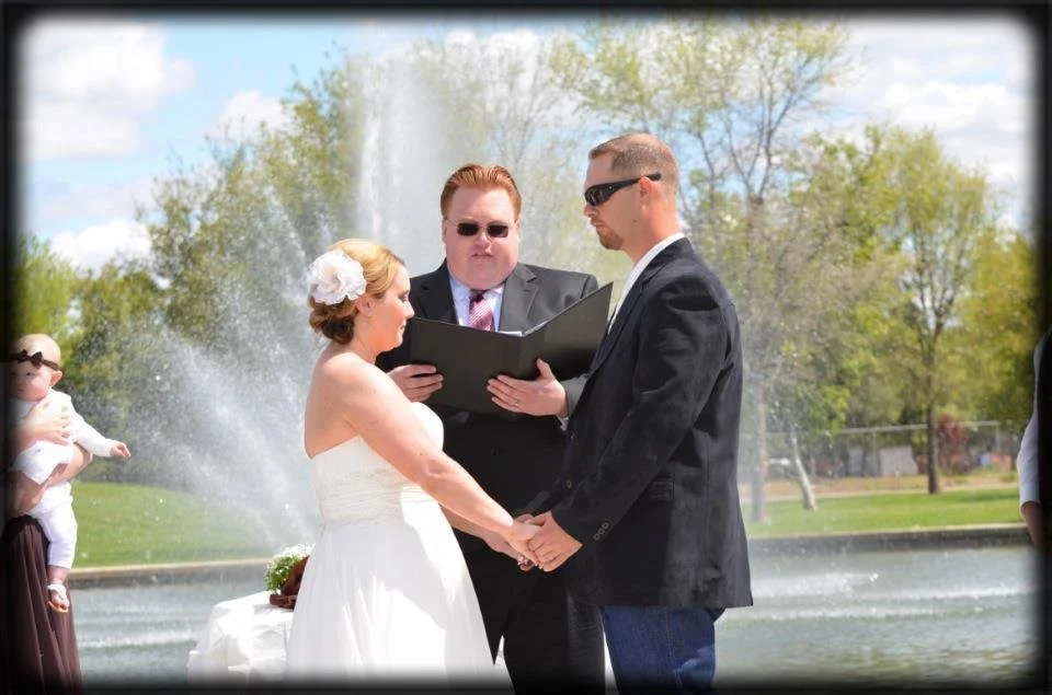 A wedding ceremony taking place outdoors near a fountain, with a bride and groom holding hands and an officiant reading vows. The bride is dressed in a white wedding gown with a large flower hair accessory, and the groom is in a black suit with sunglasses.