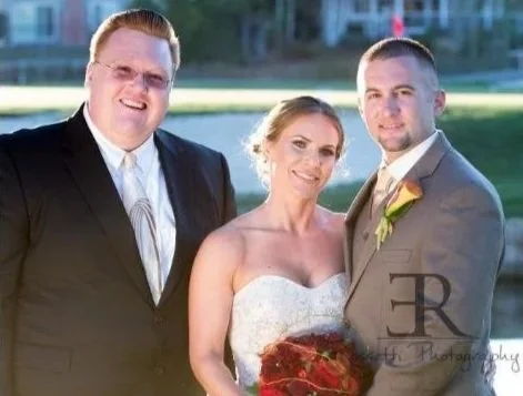 Three people dressed in formal wedding attire outdoors, with a lake and trees in the background. The woman is holding a bouquet of red flowers.