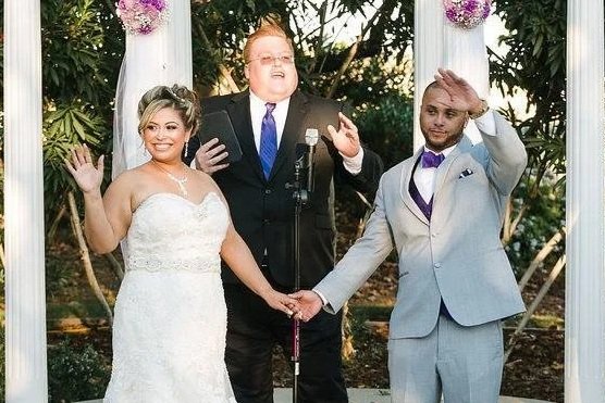 Bride and groom holding hands during wedding ceremony, with officiant behind them, outdoors with flowers and greenery.