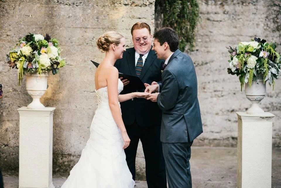 A wedding ceremony with a bride and groom holding hands, facing each other, with an officiant standing behind them, all smiling. The setting includes two large floral arrangements on pedestals, and a textured stone wall in the background.