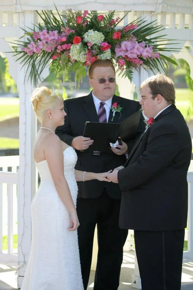 Bride and groom exchanging vows under a wedding arch decorated with pink and white flowers, officiant reading scripture