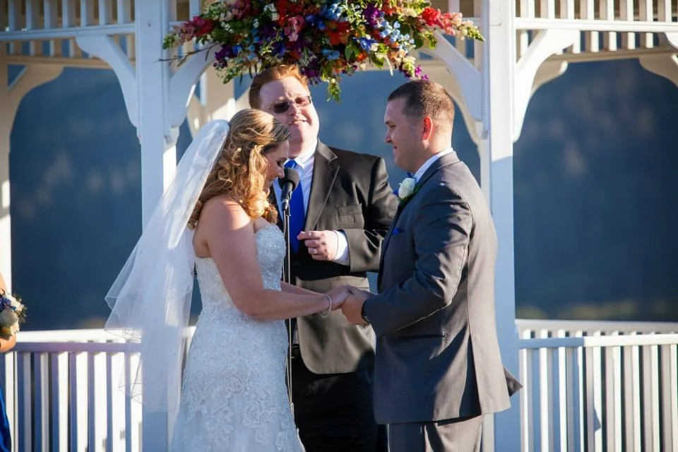 A couple getting married outdoors under a floral canopy, holding hands during their ceremony with an officiant standing behind them, in a scenic setting with a lake in the background.