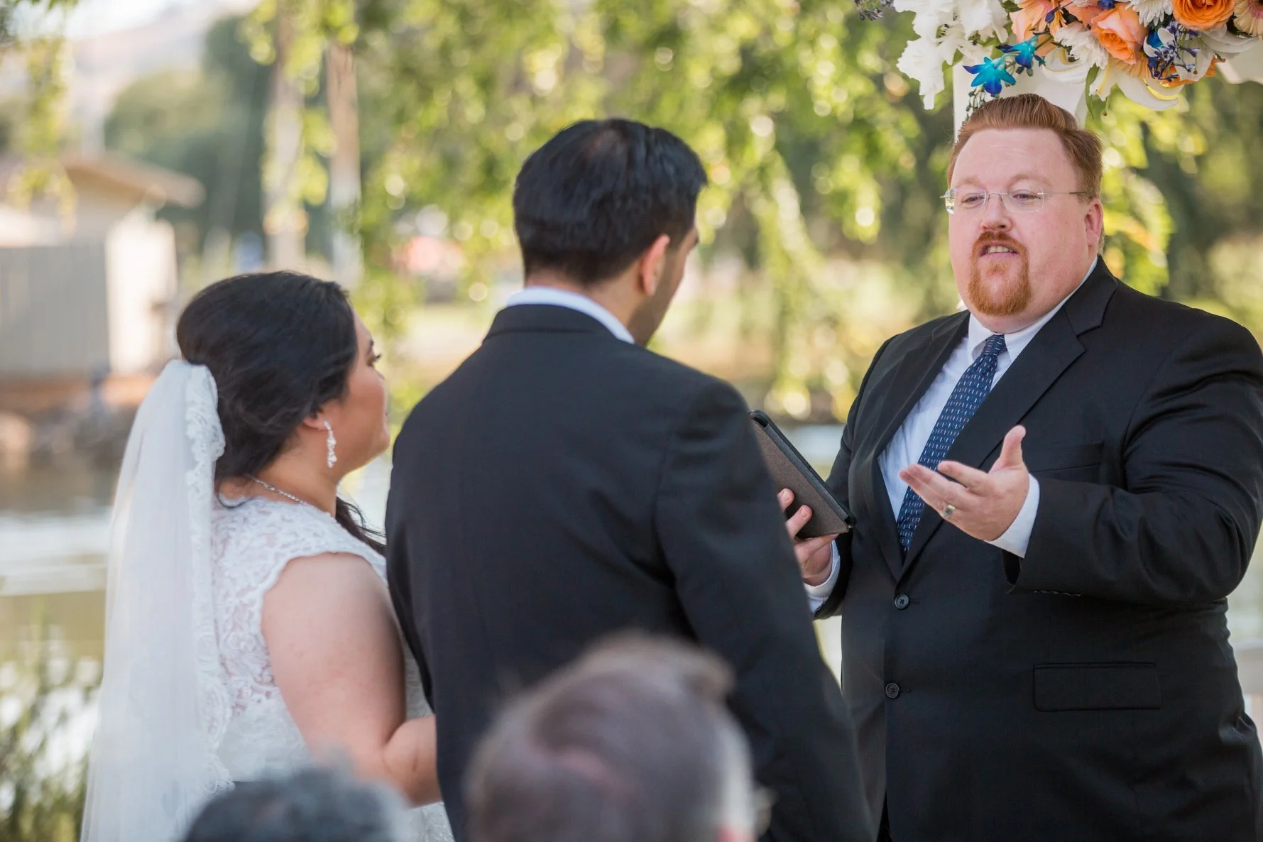 A wedding ceremony outdoors with a bride, groom, and officiant, surrounded by trees and greenery.