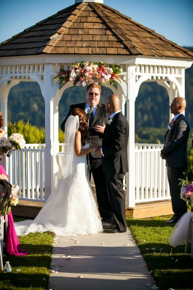 A couple exchanges vows during an outdoor wedding ceremony under a white gazebo with a flower arrangement on top, officiant present, with bridesmaids and groomsmen standing nearby.