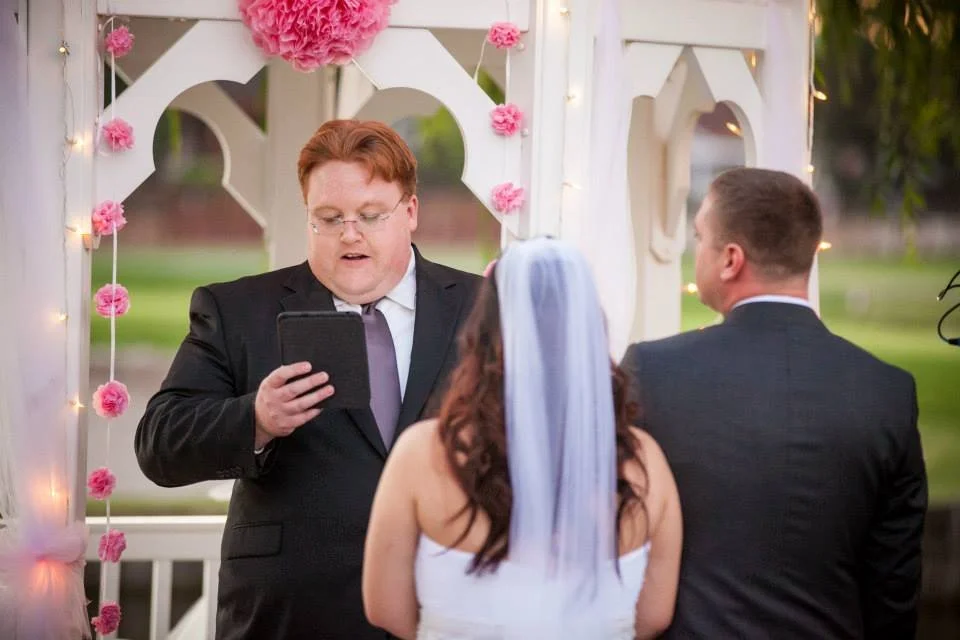 A wedding ceremony outdoors with a officiant reading vows, a bride wearing a veil and white dress, and a groom in a suit, under a decorated arch with pink flowers and string lights.