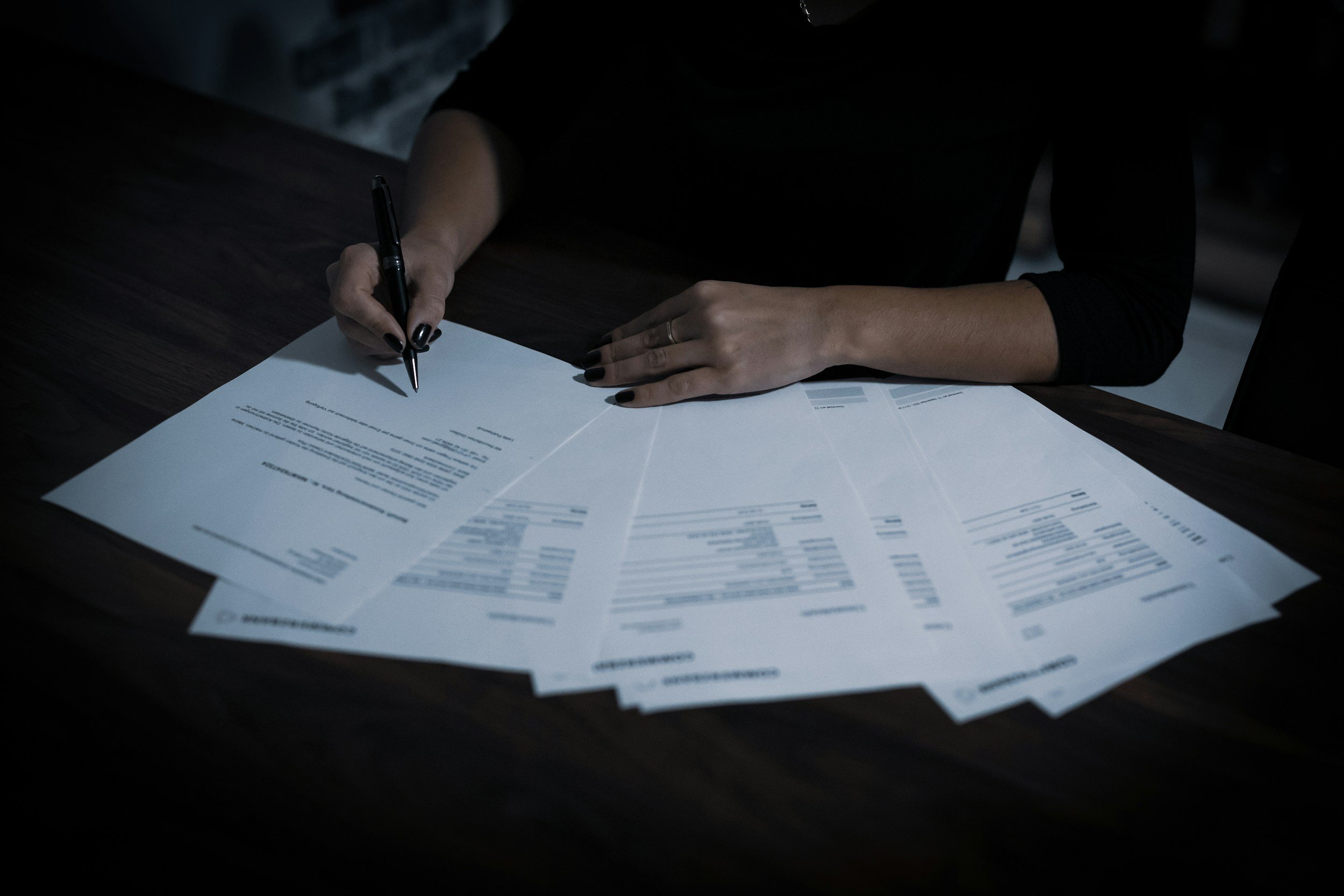 A person with black nail polish writing on a document with a black pen, several printed pages with text on a dark wooden table.