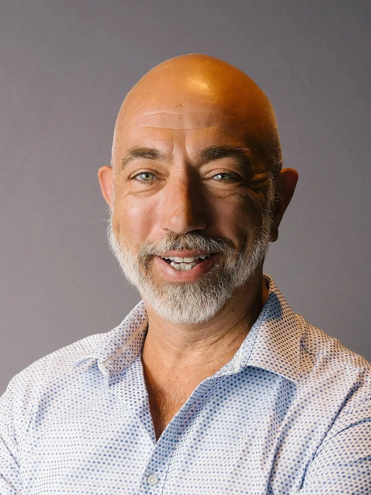 A smiling middle-aged man with a beard and light-colored eyes, bald head, wearing a light blue and white polka dot dress shirt, standing against a plain gray background.
