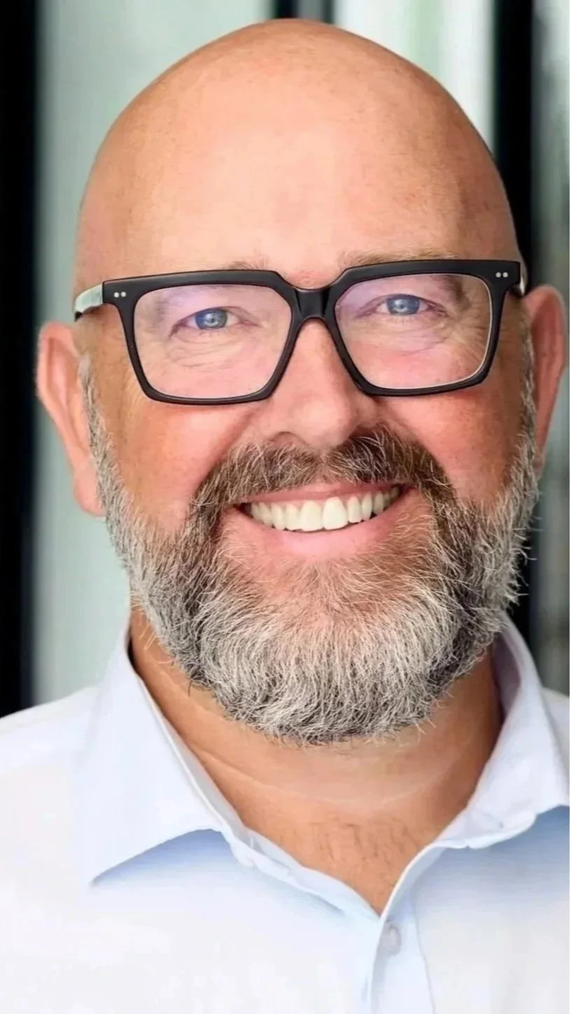A smiling bald man with glasses and a full beard, wearing a white collared shirt.